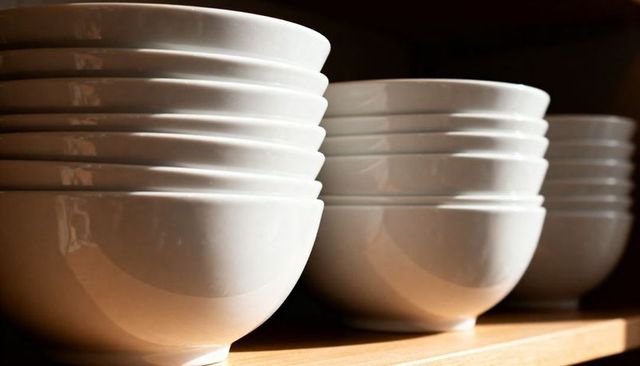 Stacked white ceramic bowls reflecting warm light on wooden shelf, glossy porcelain