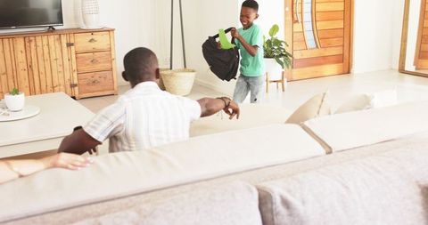 Smiling boy returning home holding backpack and notebook while parent watching from couch