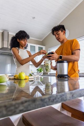 Diverse couple preparing green smoothie in modern sunlit kitchen for healthy lifestyle