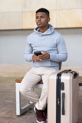 African american man waiting on urban bench holding smartphone and carry-on suitcase
