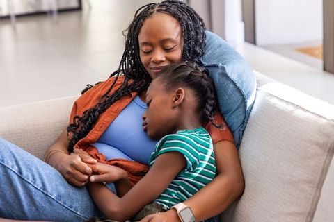 African mother and daughter resting on sofa sharing tender embrace in sunlit living room