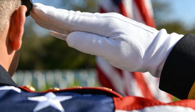 Saluting veteran wearing white glove and dark dress uniform at cemetery with american flag