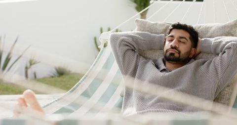 Indian man relaxing in striped hammock on patio barefoot enjoying backyard tranquility