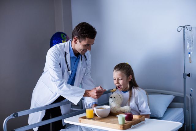 Doctor feeding breakfast to young patient in hospital room. Ideal for use in healthcare, pediatric care, medical support, and hospital recovery contexts. Highlights compassion and care in medical settings.