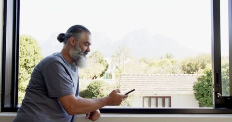 Senior Man Relaxing by Window with Smartphone, Enjoying Mountain Views