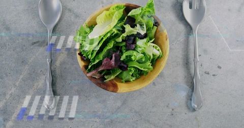 Wooden bowl holding mixed salad greens flatlay with silver spoon and fork on concrete tabletop