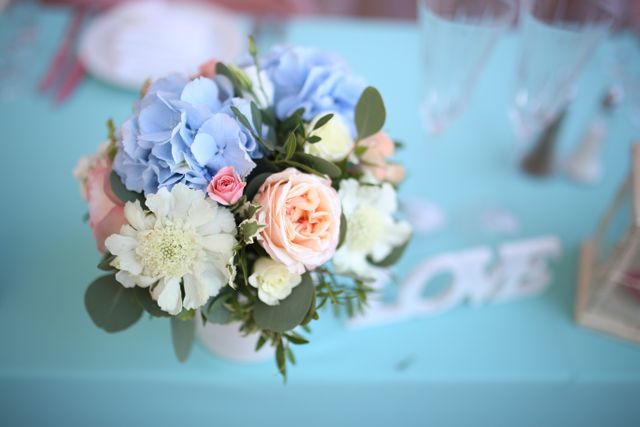 Beautiful floral centerpiece featuring a blend of pastel-colored roses, hydrangeas, and greenery arranged on a turquoise table. Ideal for use in wedding decor, romantic events, or other elegant celebrations.