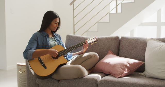 A young woman is sitting on a comfortable couch in a bright and airy living room, playing an acoustic guitar. She is casually dressed, creating a relaxed and homely atmosphere. This image is ideal for music-related websites, advertising home décor, lifestyle blogs, and promoting a relaxed and creative home environment.