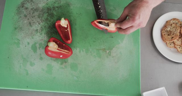 Hands visible as chef slices red bell peppers on green cutting board in professional kitchen. Bread slices shown on plate in background. Ideal for culinary tutorials, healthy eating concepts, cooking classes, recipes, and food blogs.
