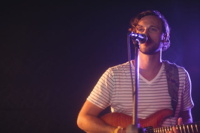 Male singer confidently performing with a guitar on stage under a spotlight. Ideal for use in articles, blogs, and promotions related to live music, concerts, musicians, and entertainment events.