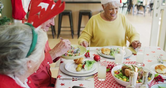 Seniors sitting around table enjoying Christmas dinner. Full plates of food, with juice, chicken legs, and salad visible. Some wear festive hats and reindeer antlers, smiling and engaging in conversation. Suitable for depicting holiday celebrations, inclusivity, and senior lifestyle.