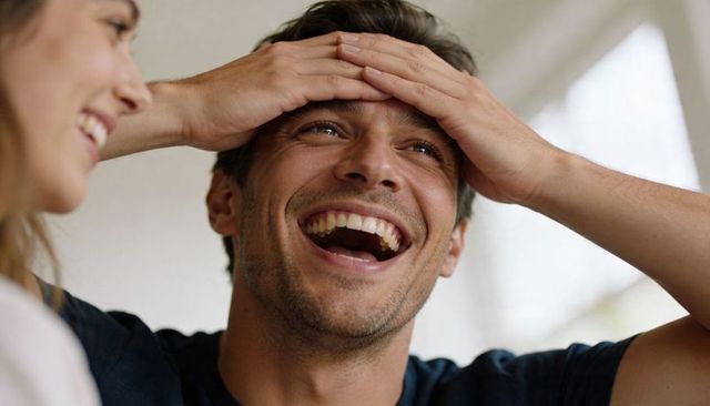Young man laughing holding head while partner smiling nearby in bright sunlit living room