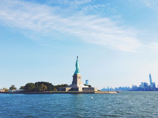 The Statue of Liberty stands tall against a clear blue sky with the New York City skyline as a beautiful backdrop. Perfect to use for travel guides, tourism promotion, history documentaries, and educational materials highlighting iconic landmarks.