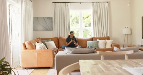 African american woman relaxing on tan sofa using smartphone and wired headphones