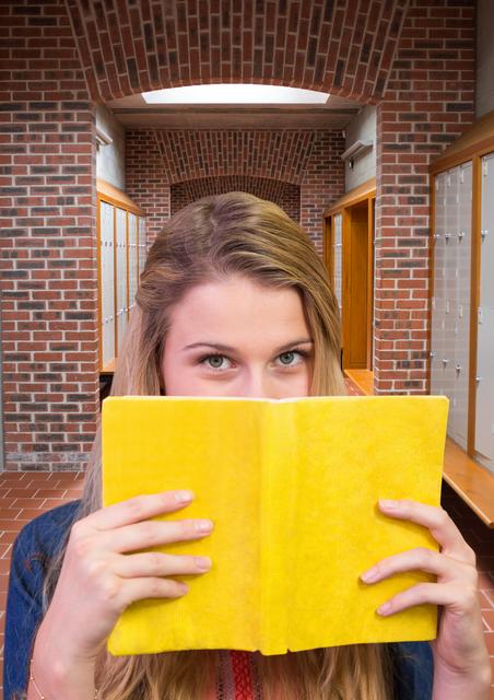 Young female student holding yellow book close to her face, concealing identity while standing in school locker room. With the backdrop of brick walls, lockers, and benches, this image is great for depicting themes of education, privacy, adolescence, and student life in academic environments.