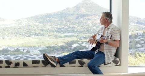 Senior man playing acoustic guitar on window bench overlooking valley and mountain view