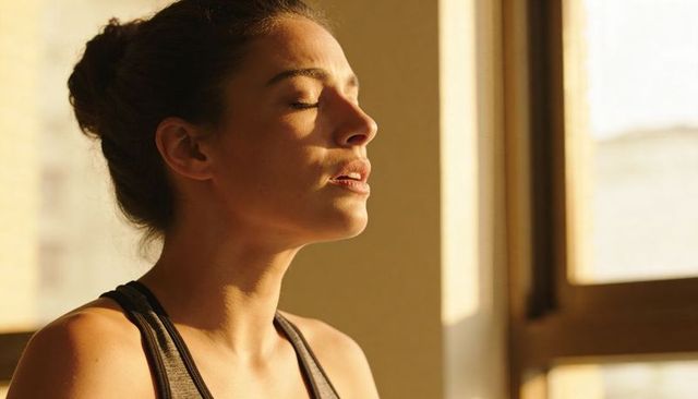 Woman meditating in warm morning sunlight by window, eyes closed, minimalist wellness headshot
