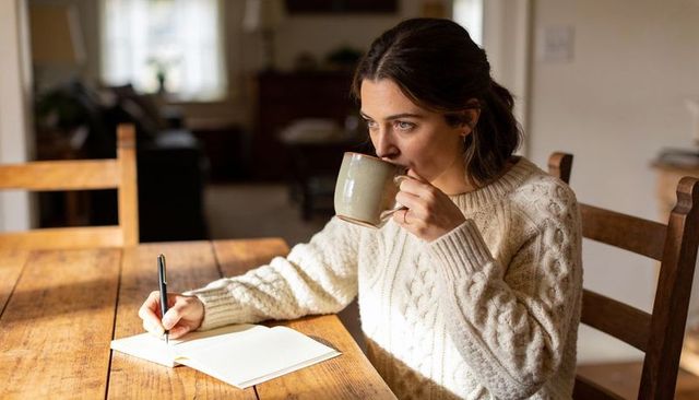 Sipping coffee and writing in journal at sunlit rustic dining table, cozy knit sweater