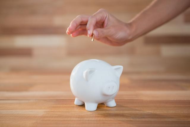 Hand inserting coin into white piggy bank on wooden surface. Ideal for illustrating concepts related to saving money, personal finance, budgeting, and financial planning. Suitable for use in financial blogs, educational materials, and advertisements promoting savings and investments.