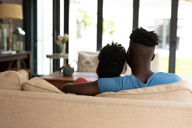 Young African American couple sitting on a sofa with the man's arm around the woman, enjoying a quiet moment together in a modern living room. Ideal for use in lifestyle blogs, relationship advice articles, home decor websites, and advertisements promoting comfortable living spaces.