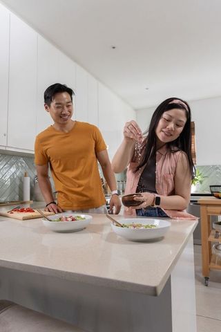 Asian couple cooking together on quartz island while woman sprinkles seasoning into salad bowls
