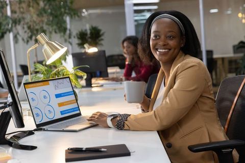 Smiling professional woman holding mug and working on laptop with business analytics