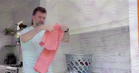 Mature man folding coral t-shirt in bright kitchen with laundry basket