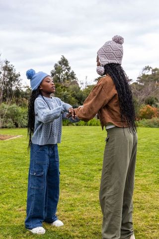 African american woman holding hands with girl playing on park lawn in knit hats
