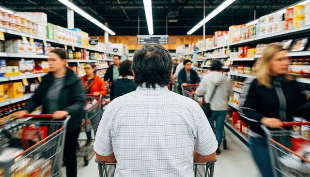 Senior man pushing shopping cart through crowded supermarket aisle, busy grocery shoppers