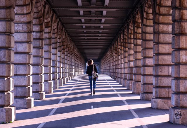 Person walking through an urban bridge tunnel filled with repeated stone columns and sunlit shadows, capturing symmetry and perspective. Sun shines through casting long shadows creating visual depth. Would work well for designs needing themes of journey, urban exploration, solitude, or architectural details.