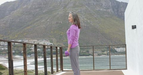 Mature woman exercising on coastal balcony holding purple dumbbell, mountain and sea view