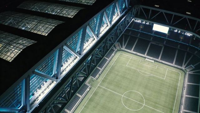 Aerial stadium illuminating empty soccer pitch at night with modern truss roof and floodlights