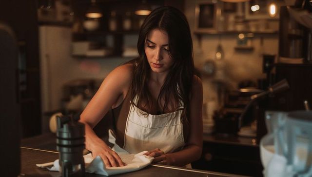 Young female barista cleaning coffee counter in warm cafe light wearing apron and tamper