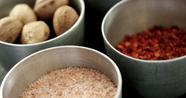Three metal bowls contain various cooking ingredients: whole nutmegs, coarse pink salt, and crushed red pepper flakes. These spices and seasonings are essential for adding flavor and depth to a wide range of culinary dishes.