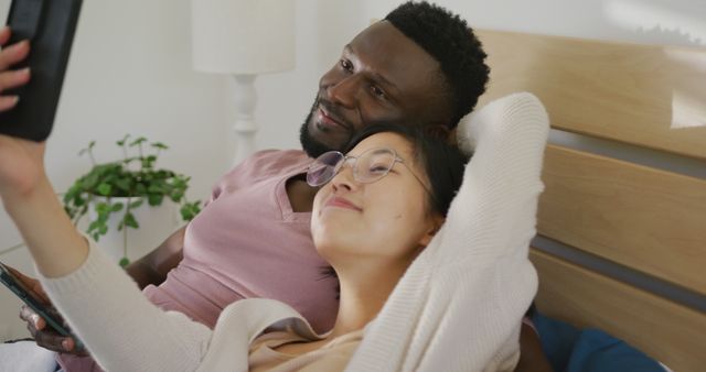 Happy diverse couple using smartphone and lying in bedroom. Spending quality time at home concept.