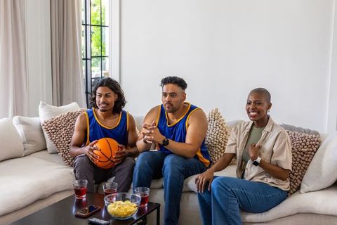 Diverse friends watching basketball game at home on couch wearing team jerseys