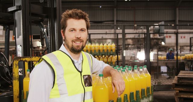 Confident worker wearing safety vest standing by production line with bottles of orange juice, smiling at camera. Perfect for content about industrial manufacturing, production management, factory operations, employee safety, quality control, and beverage industry.