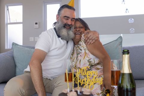 Mature couple celebrating birthday at home hugging on sofa with cake and champagne