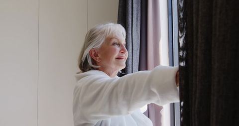 Senior woman in bathrobe stretching curtains by window