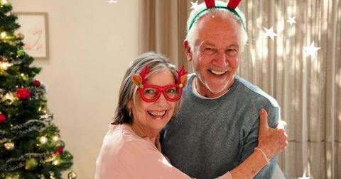 Senior couple embracing by christmas tree wearing reindeer glasses and antler headband