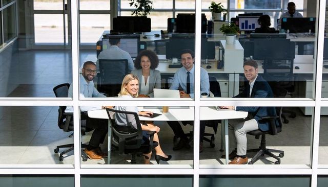 Business team discussing strategy in glass-walled open-plan office with laptops and coffee