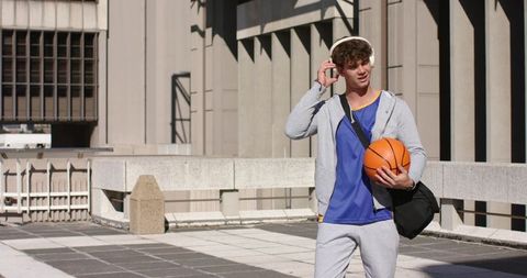 Young man holding basketball on sunlit urban rooftop wearing headphones and gym bag