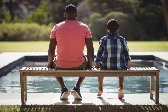 Father and son sitting together on a bench by a pool, enjoying a relaxing moment outdoors. Ideal for use in family-oriented content, parenting blogs, advertisements promoting family activities, or articles about father-son relationships and bonding.