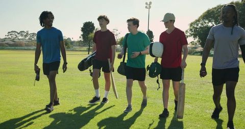 Multicultural cricket teammates walking to practice carrying bats and helmet on park grass