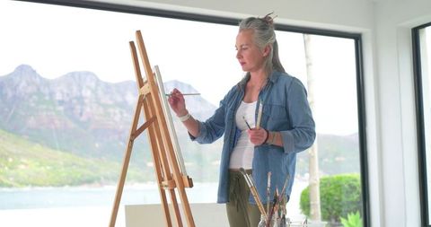 Senior Woman Painting at Easel by Waterfront in Sunlit Studio with Mountain View, Art Supplies