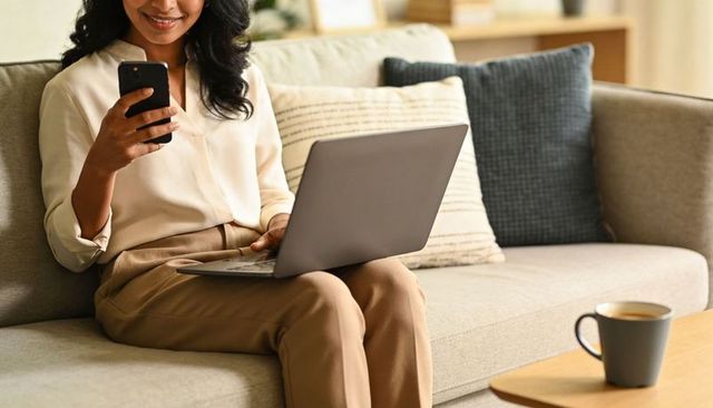 Woman using smartphone and laptop on sofa, checking messages and working remotely at home
