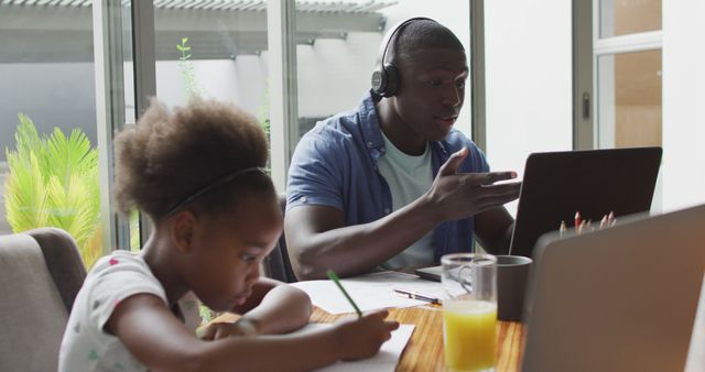 Father working on laptop while wearing headset, daughter doing homework at the same table. Great to illustrate work-life balance, remote work setups, family bonding, and modern parenting. Useful for promoting home offices, online education tools, and family-friendly policies.