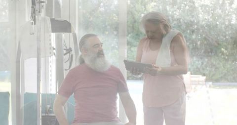 Mature couple exercising with tablet in bright home gym overlooking patio and pool