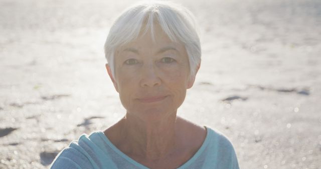 Senior woman with grey hair, dressed in a light casual shirt, captured in evening light on a tranquil beach. Her serene and thoughtful expression evokes calm and peaceful moments by the sea. Great for use in materials related to aging gracefully, beach lifestyle, retirement living, personal reflections, or mental well-being.