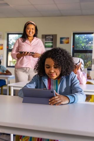 Primary student using tablet with folio stand while teacher supervising digital classroom